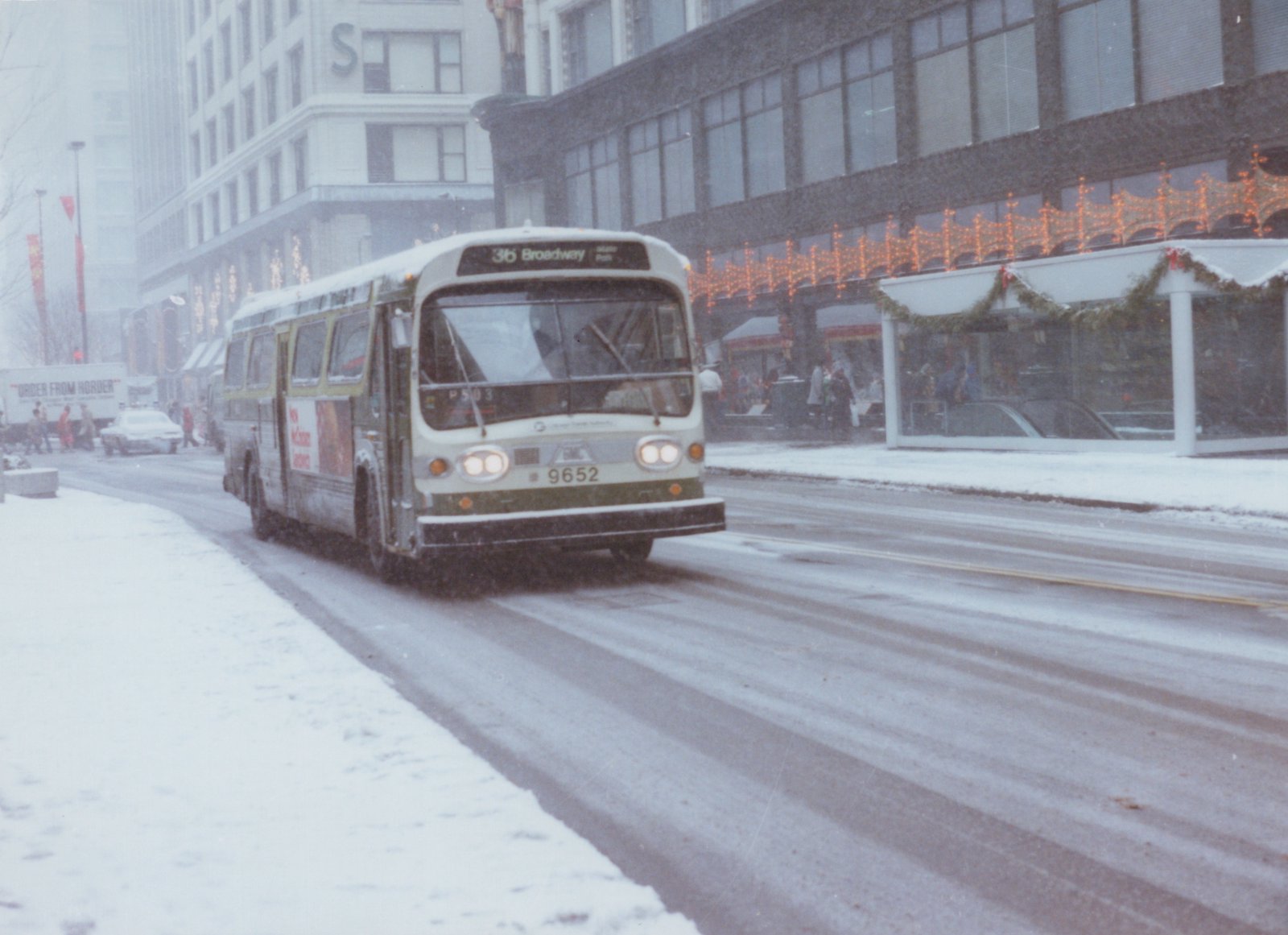 A green and ivory CTA bus drives down a snow-covered city street, with holiday lights and pedestrians visible on the sidewalk and a large building with an 'S' sign in the background.