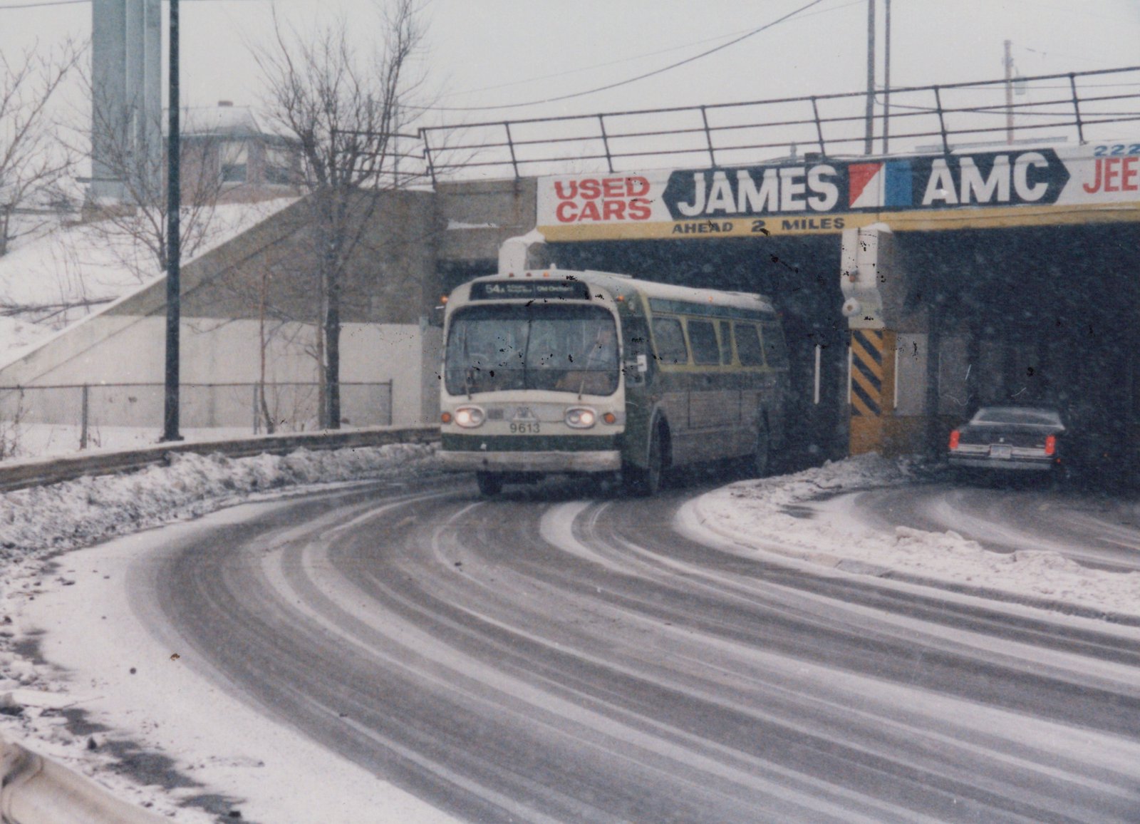 A green and cream CTA bus with the number 9613 drives on a snowy, curving road under a bridge with a 'USED CARS JAMES AMC' sign.