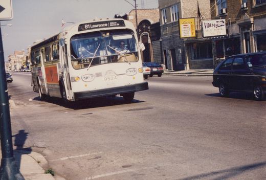 A white CTA bus with green and orange stripes, number 9524, turns a corner on a sunny day, with storefronts and cars in the background.