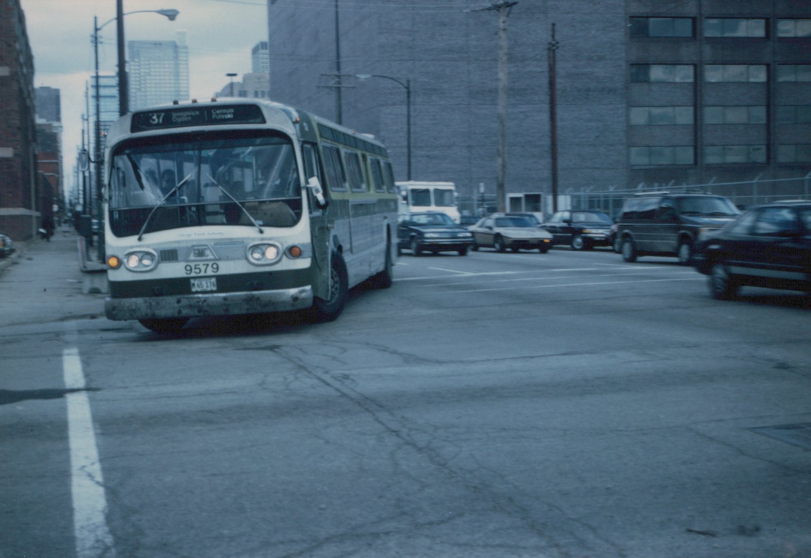 A white and green CTA bus with the number 9579 on the front is stopped on a city street, with several cars visible in the background and a large brick building on the right.