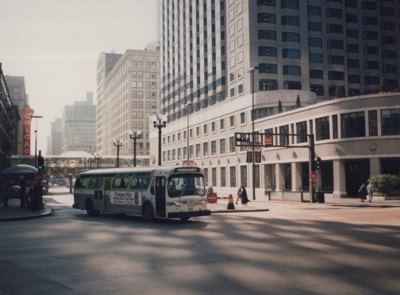 A white and green CTA bus with the number 9506 on its front drives through a city street with tall buildings and pedestrians in the background.