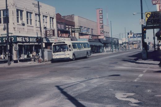A white and green CTA bus numbered 9472 is stopped at an intersection on a sunny day, with storefronts including Mel's Men's Shop and a tall sign for 'ANNES' in the background.