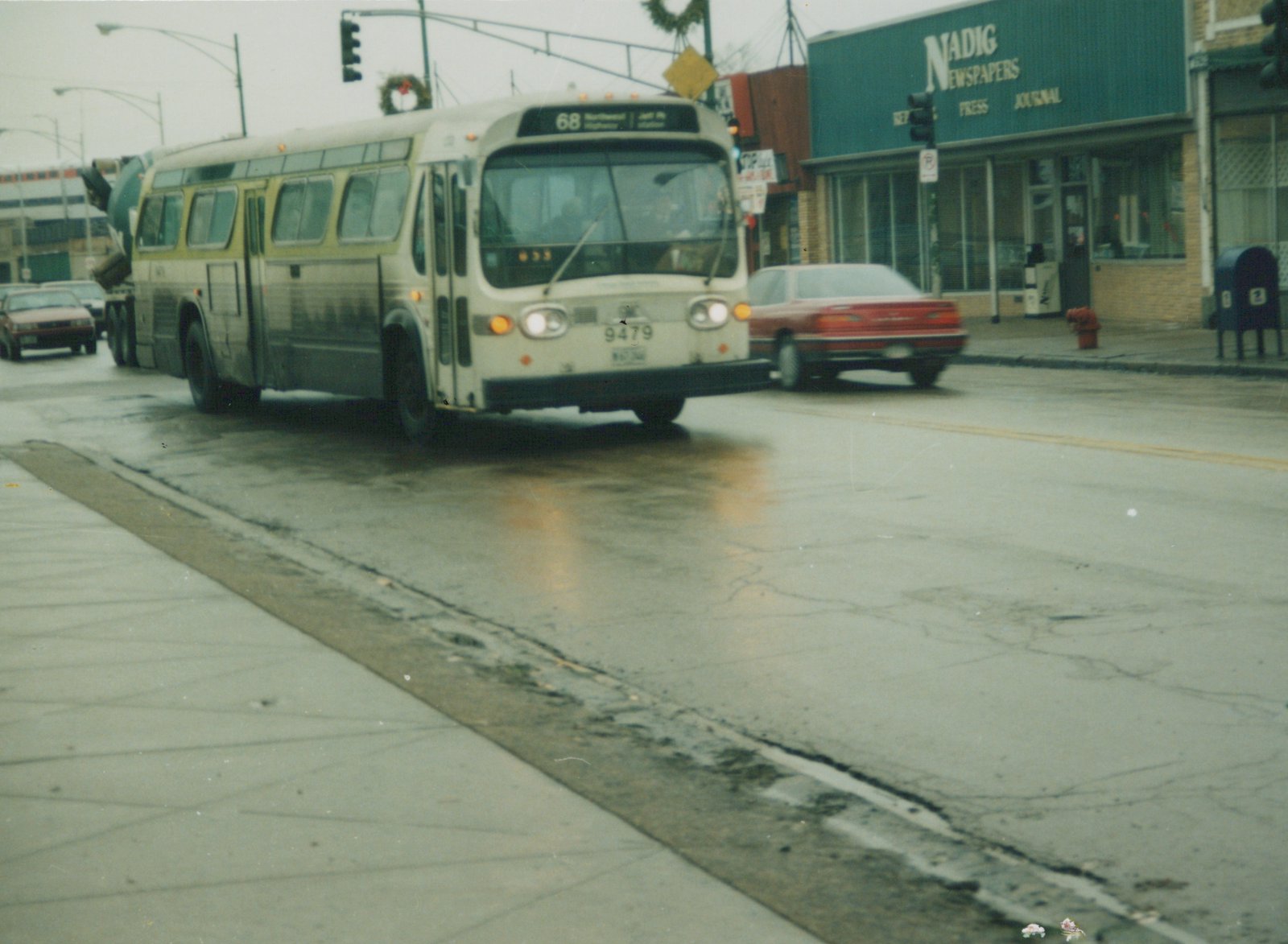 A white and green CTA bus numbered 9479 travels down a wet street, with a red car and storefronts in the background.