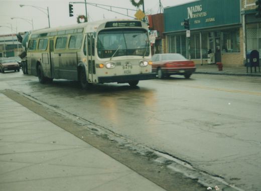 A white and green CTA bus numbered 9479 travels down a wet street, with a red car and storefronts in the background.