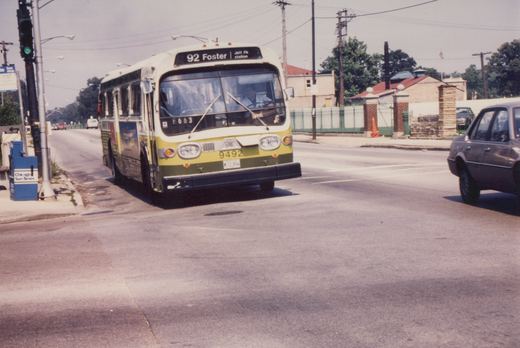 A green and cream CTA bus is turning at an intersection on a sunny day, with a newsbox and a cemetery sign visible in the background.