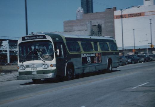 A green and cream CTA bus with fleet number 9398 is driving down a city street, with a '12 Roosevelt Monitor' destination sign visible on the front and a McDonald's ad on the side.