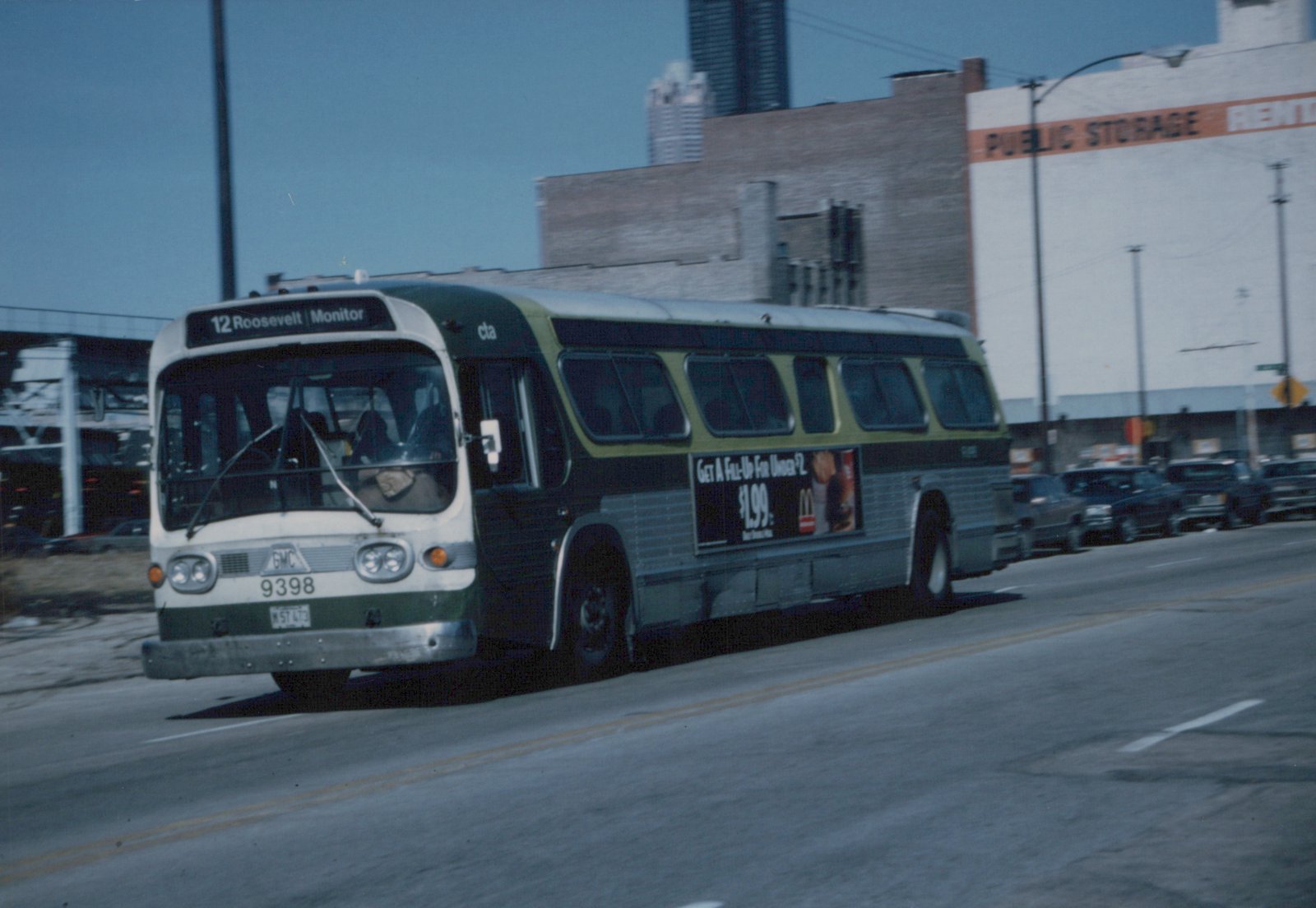 A green and cream CTA bus with fleet number 9398 is driving down a city street, with a '12 Roosevelt Monitor' destination sign visible on the front and a McDonald's ad on the side.