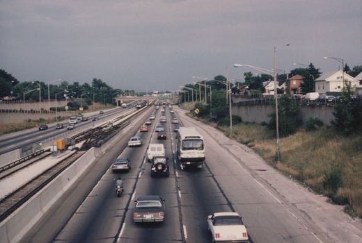 A white CTA bus with route 61 Lawrence travels on a multi-lane highway next to a railway track with a train, surrounded by cars and a motorcycle under a cloudy sky.