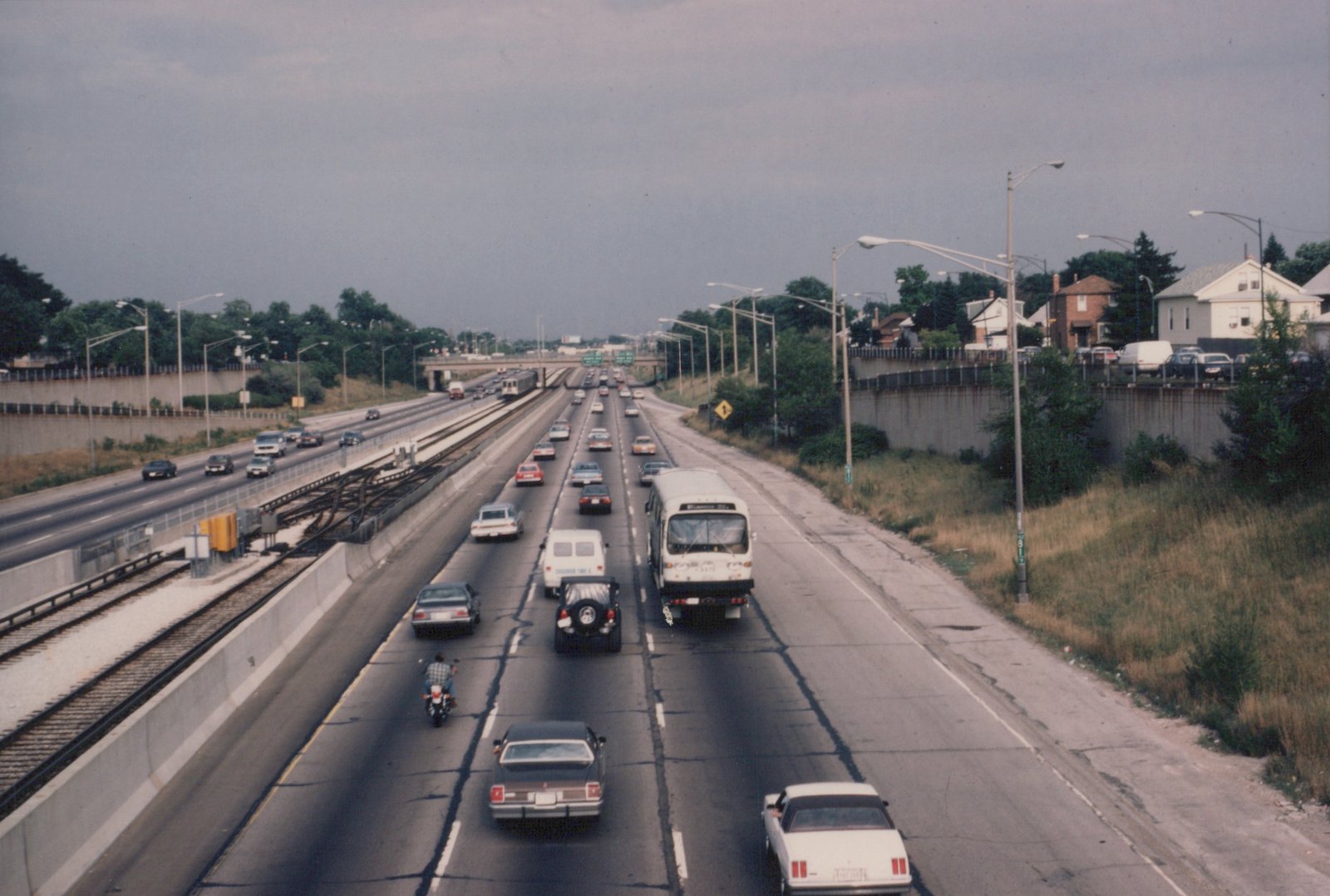 A white CTA bus with route 61 Lawrence travels on a multi-lane highway next to a railway track with a train, surrounded by cars and a motorcycle under a cloudy sky.