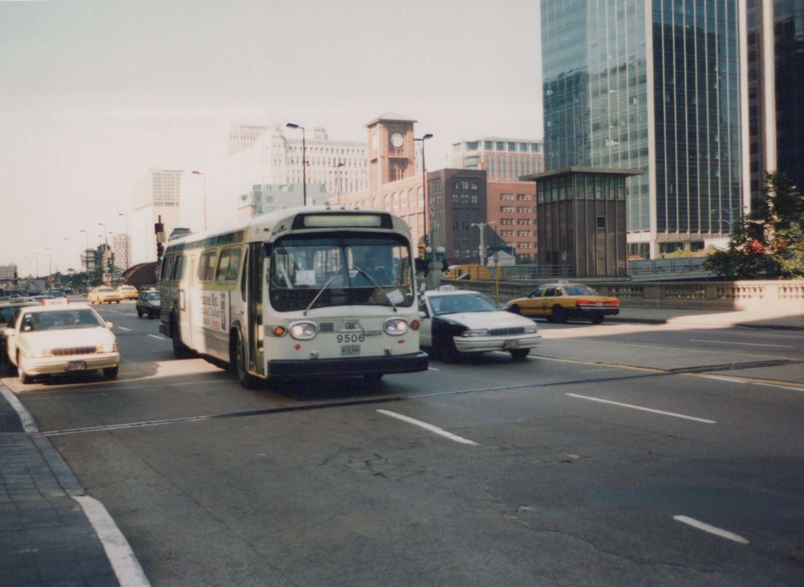 A white and green Chicago Transit Authority bus with the number 9506 is driving on a multi-lane city street surrounded by taxis and tall buildings.