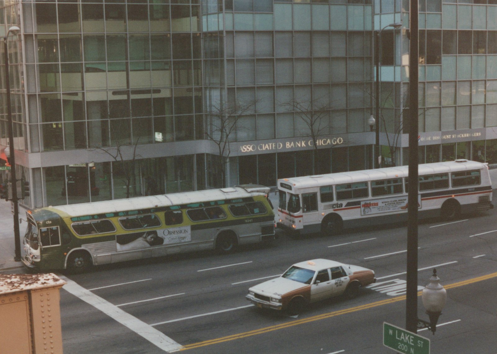 A high-angle photo captures two CTA buses, one green and white and one white with a red stripe, on a city street. A brown and white taxi is in the foreground, and a large glass office building with an 'Associated Bank Chicago' sign is behind them.