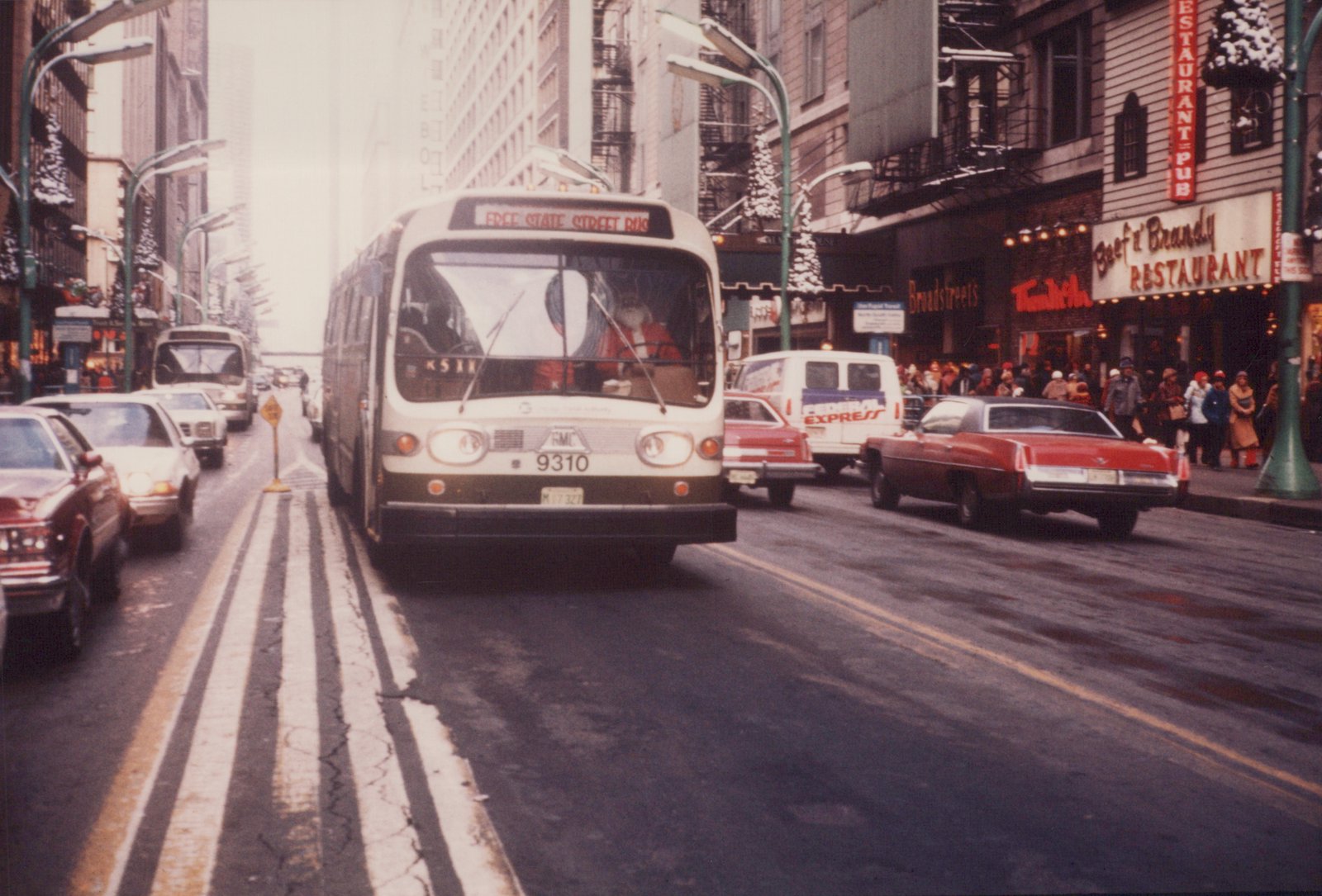 Front view of a white and black Chicago Transit Authority bus with the number 9310, driven by a man dressed as Santa Claus, on a city street lined with cars and decorated for Christmas.