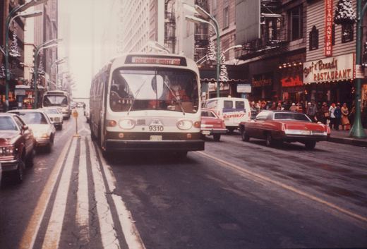 Front view of a white and black Chicago Transit Authority bus with the number 9310, driven by a man dressed as Santa Claus, on a city street lined with cars and decorated for Christmas.