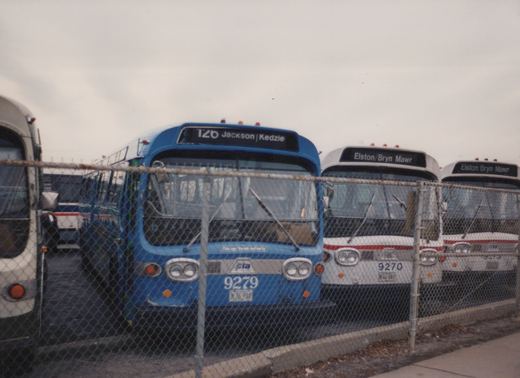 A row of Chicago Transit Authority buses, with a blue bus numbered 9279 and two white buses numbered 9270 and 9273, are parked behind a chain-link fence on an overcast day.