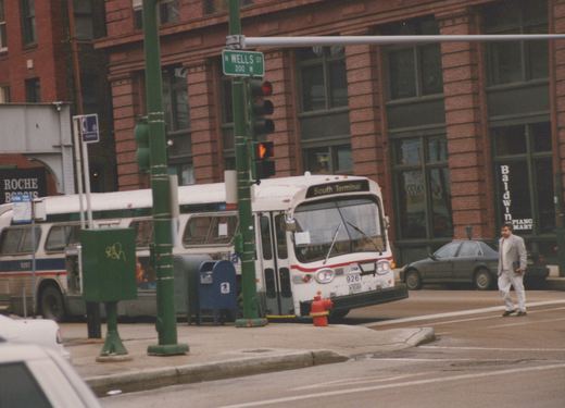 A white CTA bus with red and blue stripes, numbered 9267, is stopped at a street intersection in Chicago, with a pedestrian crossing the street and a second bus visible behind it.