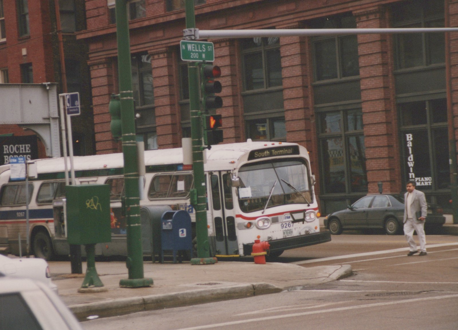 A white CTA bus with red and blue stripes, numbered 9267, is stopped at a street intersection in Chicago, with a pedestrian crossing the street and a second bus visible behind it.
