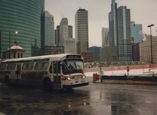 A white and red CTA bus, number 9267, drives on a rain-slicked street with tall buildings behind it.