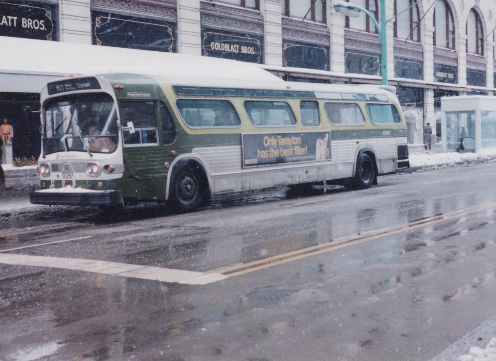 A green and cream Chicago Transit Authority bus is parked on a snowy city street, with a storefront and a bus shelter in the background.