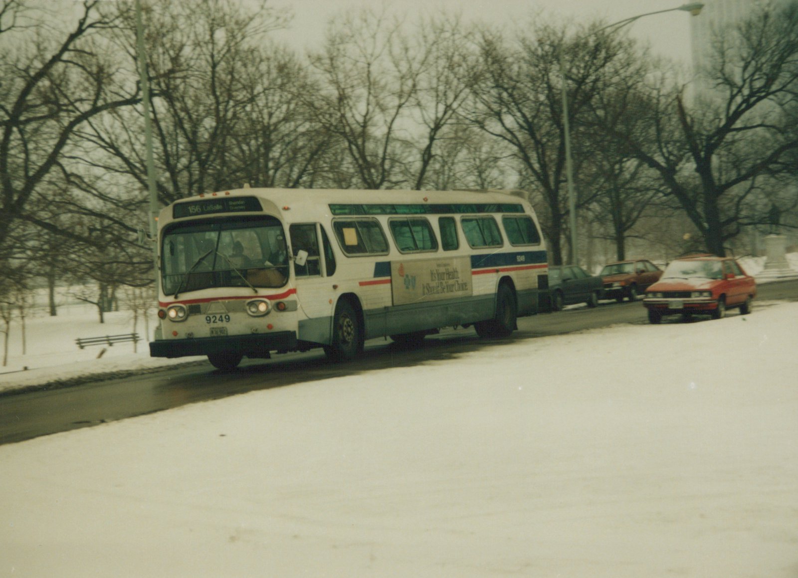 A white and blue Chicago Transit Authority bus, number 9249, drives down a snow-covered street with bare trees in the background.