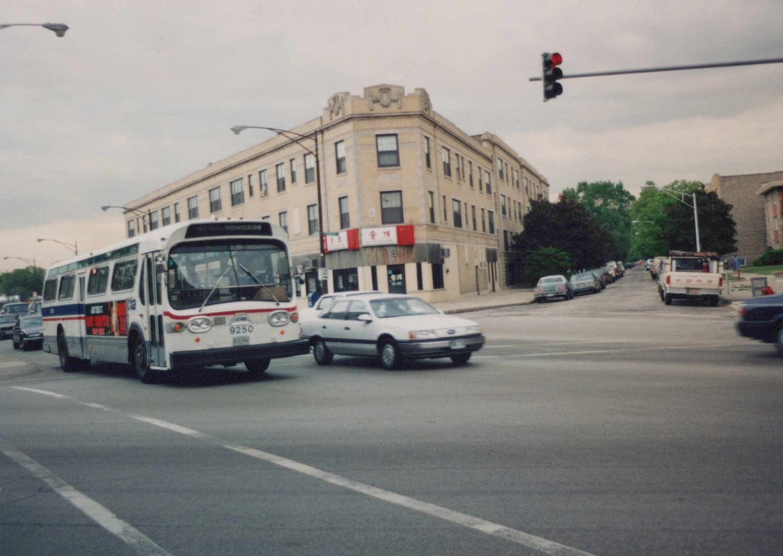 A white and blue CTA bus, number 9250, is at a street corner with a red traffic light, surrounded by cars and a corner building with storefronts.