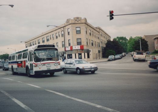 A white and blue CTA bus, number 9250, is at a street corner with a red traffic light, surrounded by cars and a corner building with storefronts.