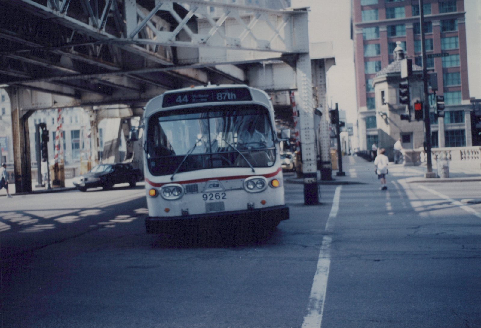 A white CTA bus with fleet number 9262 is driving on a city street under an elevated railroad, with pedestrians on the sidewalk.