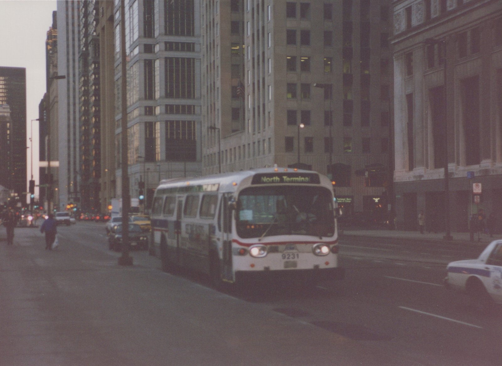 A white, red, and blue CTA bus with the number 9231 and destination 'North Terminal' is driving down a city street lined with skyscrapers at dusk, with pedestrians and other vehicles visible.
