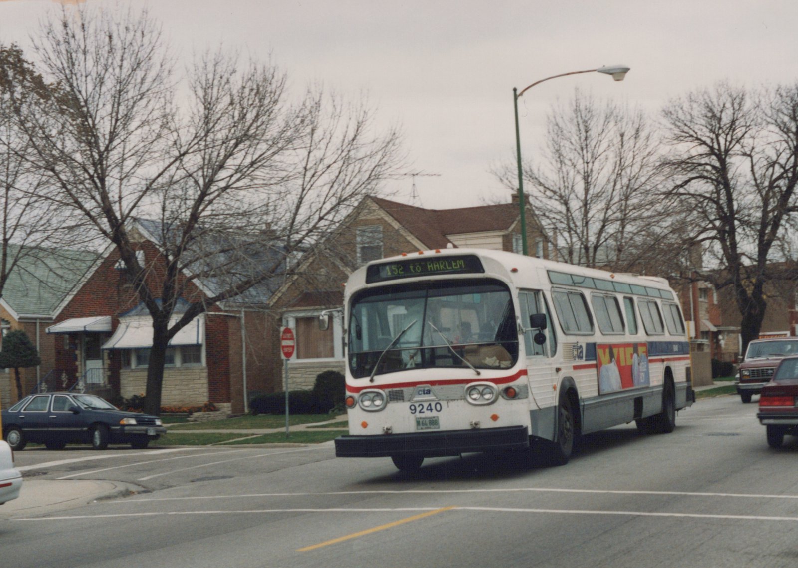 A white CTA bus with a red stripe, number 9240, is driving down a street lined with bare trees and houses; the destination sign reads '152 to HARLEM'.