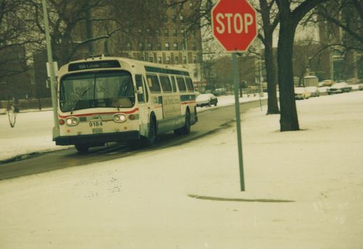 An older white and red CTA bus drives down a snow-covered street next to a stop sign, with apartment buildings in the background.