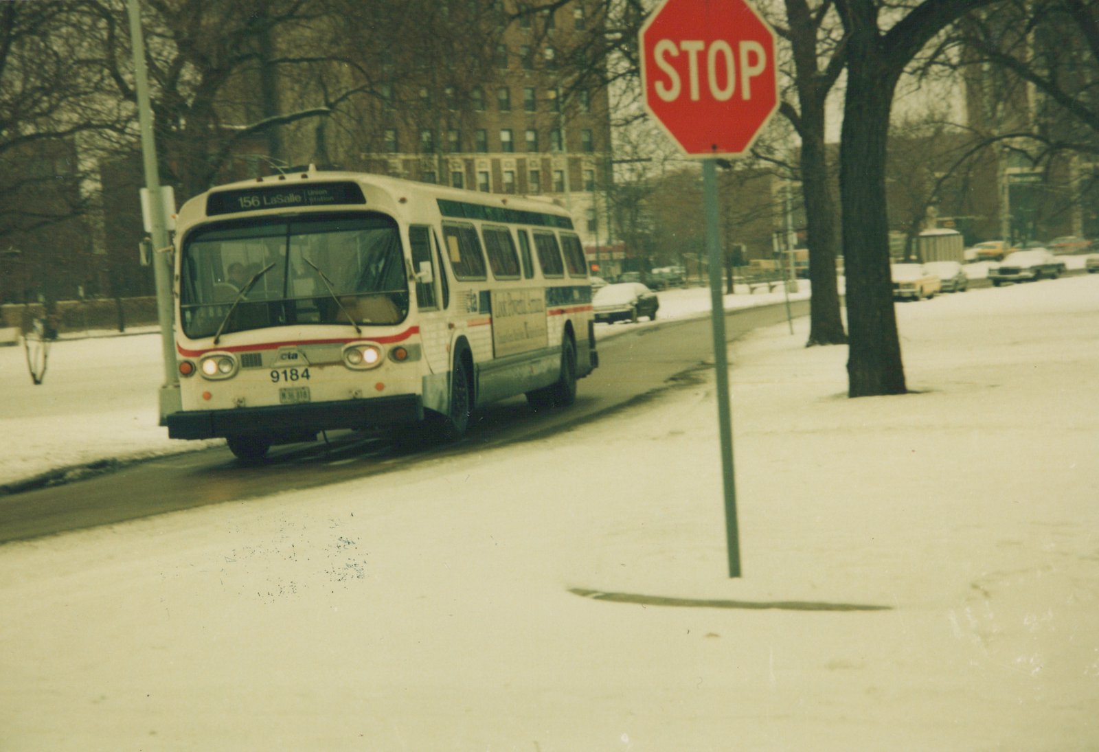 An older white and red CTA bus drives down a snow-covered street next to a stop sign, with apartment buildings in the background.