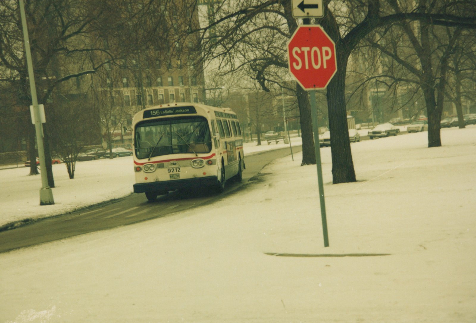 A white CTA bus with a red stripe, number 9212, drives down a snow-covered street with bare trees and a stop sign nearby.