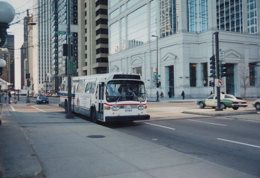 A white and red CTA bus with the number 9194 is stopped on a city street with tall buildings in the background and a green and white taxi nearby.