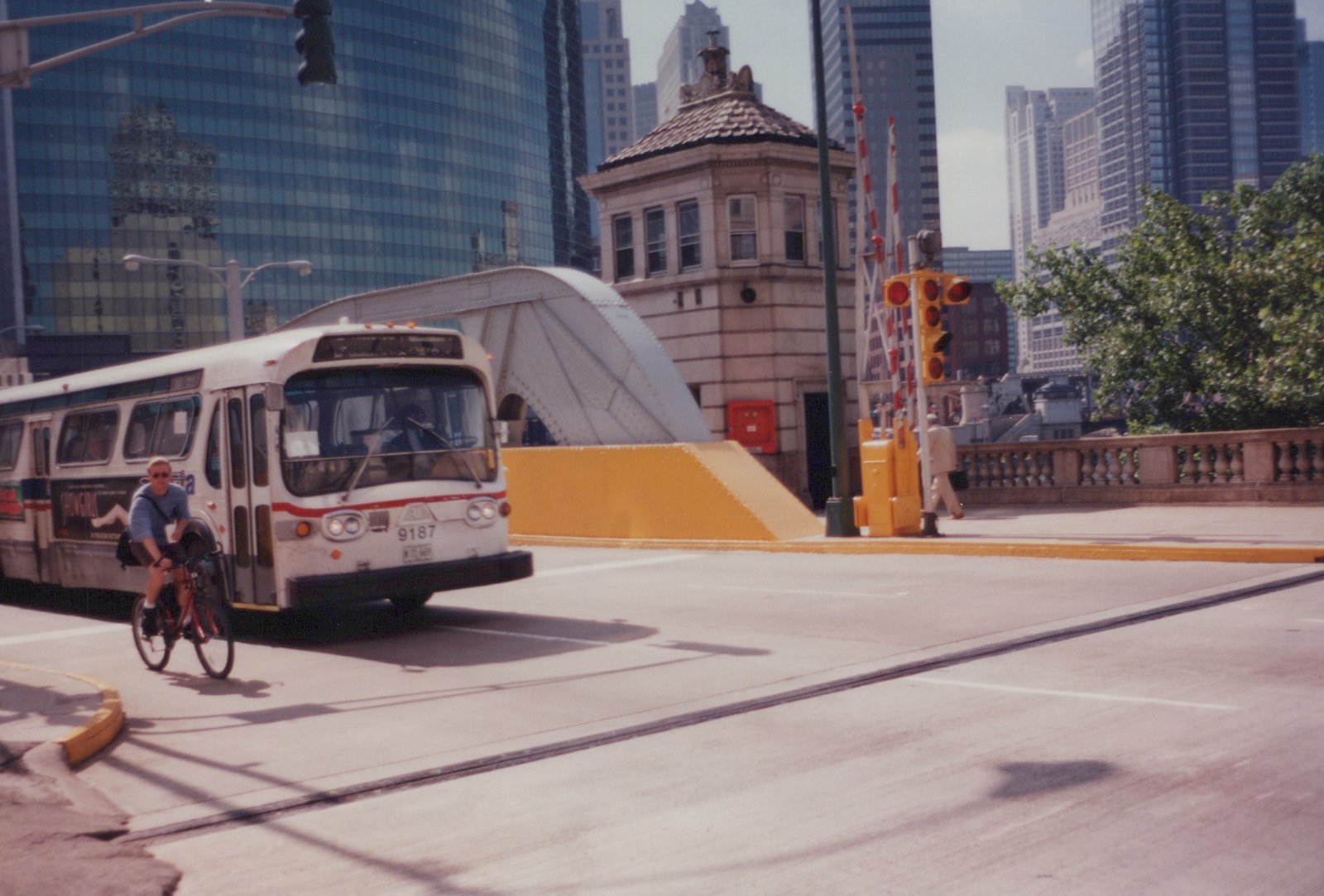 A white and red CTA bus with the number 9187 on its front drives across a bridge in a city, with a person on a bicycle in front of it and tall buildings behind.