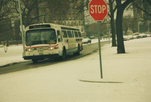 A white CTA bus with the number 9184 and destination '156 LaSalle' is driving on a snow-covered street next to a red stop sign; bare trees and parked cars are in the background.