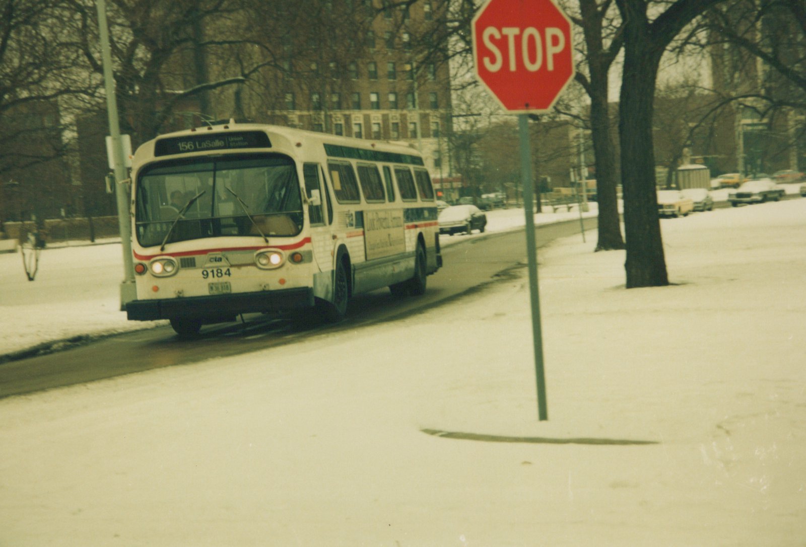A white CTA bus with the number 9184 and destination '156 LaSalle' is driving on a snow-covered street next to a red stop sign; bare trees and parked cars are in the background.