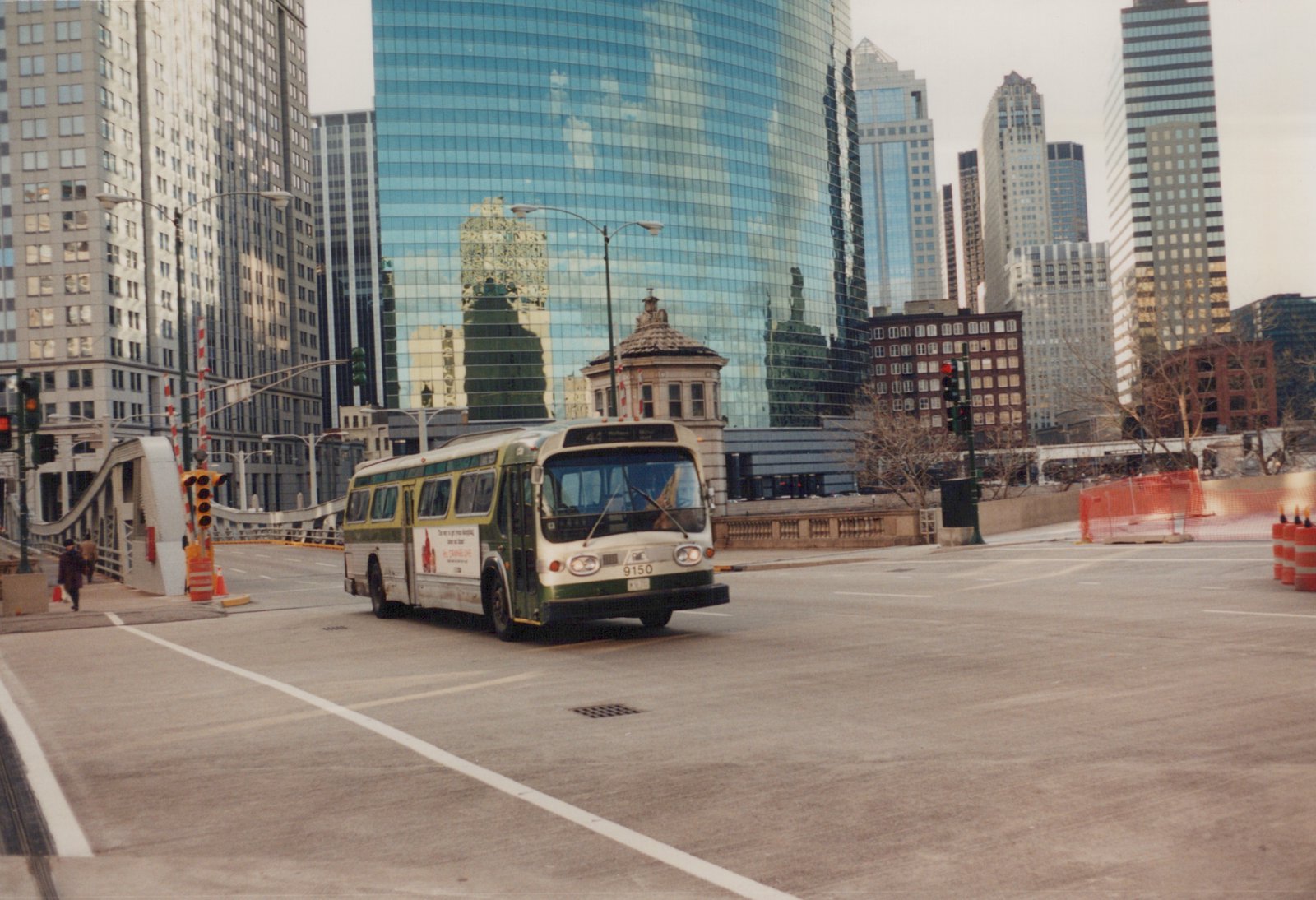 A green and cream CTA motor bus with the number 9150 on its front is driving on a city street with tall skyscrapers in the background.