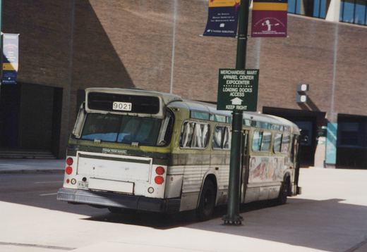 A weathered green and white city bus, seen from the rear corner, stands at a curb by a brick building, beside a green lamp post with a sign for the Merchandise Mart, Apparel Center and Expocenter loading docks.