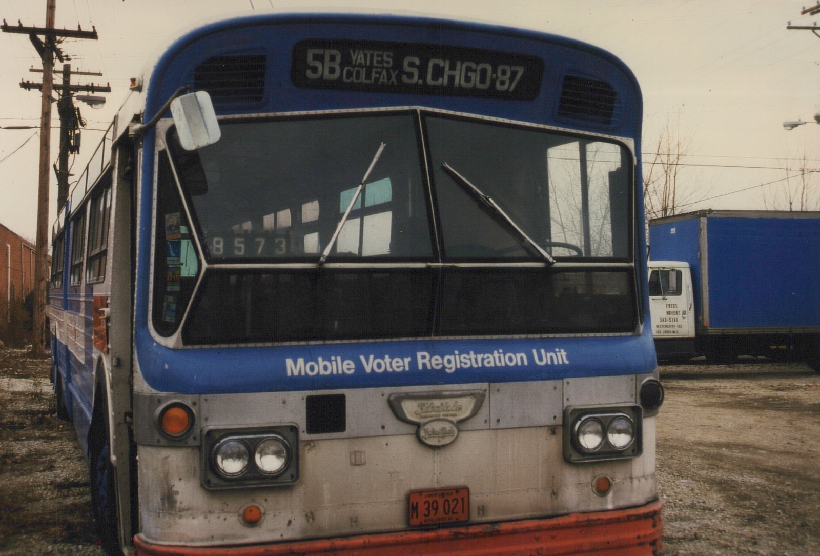 Front view of a blue and white CTA bus with 'Mobile Voter Registration Unit' lettered on its front, parked in a yard with a truck behind it.