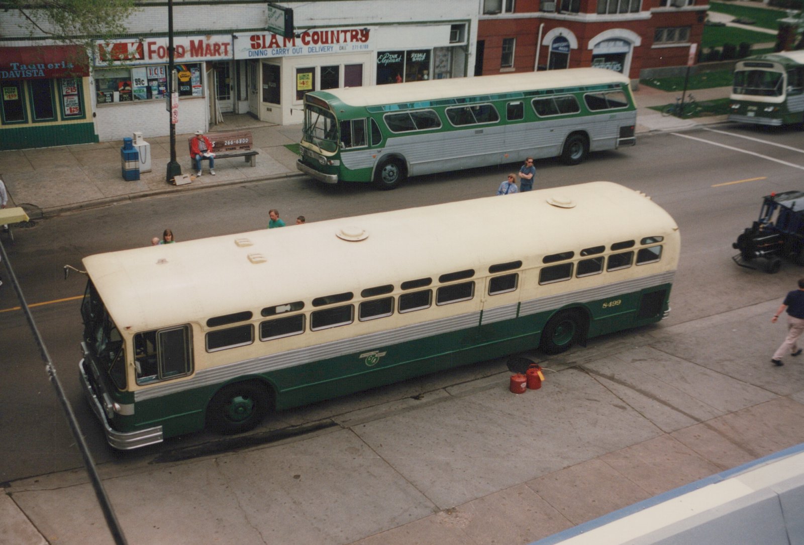 An elevated view of two green and cream CTA buses, numbered 8499 and 801, on a city street with people standing nearby and storefronts including a 'Siam Country' restaurant.