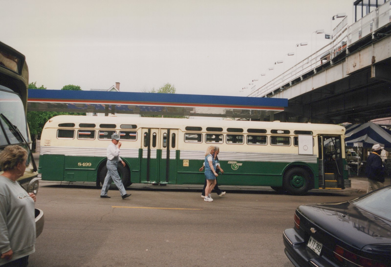 Side view of a green and cream CTA bus with number 8499, parked under a steel elevated railway with people walking by on a cloudy day.