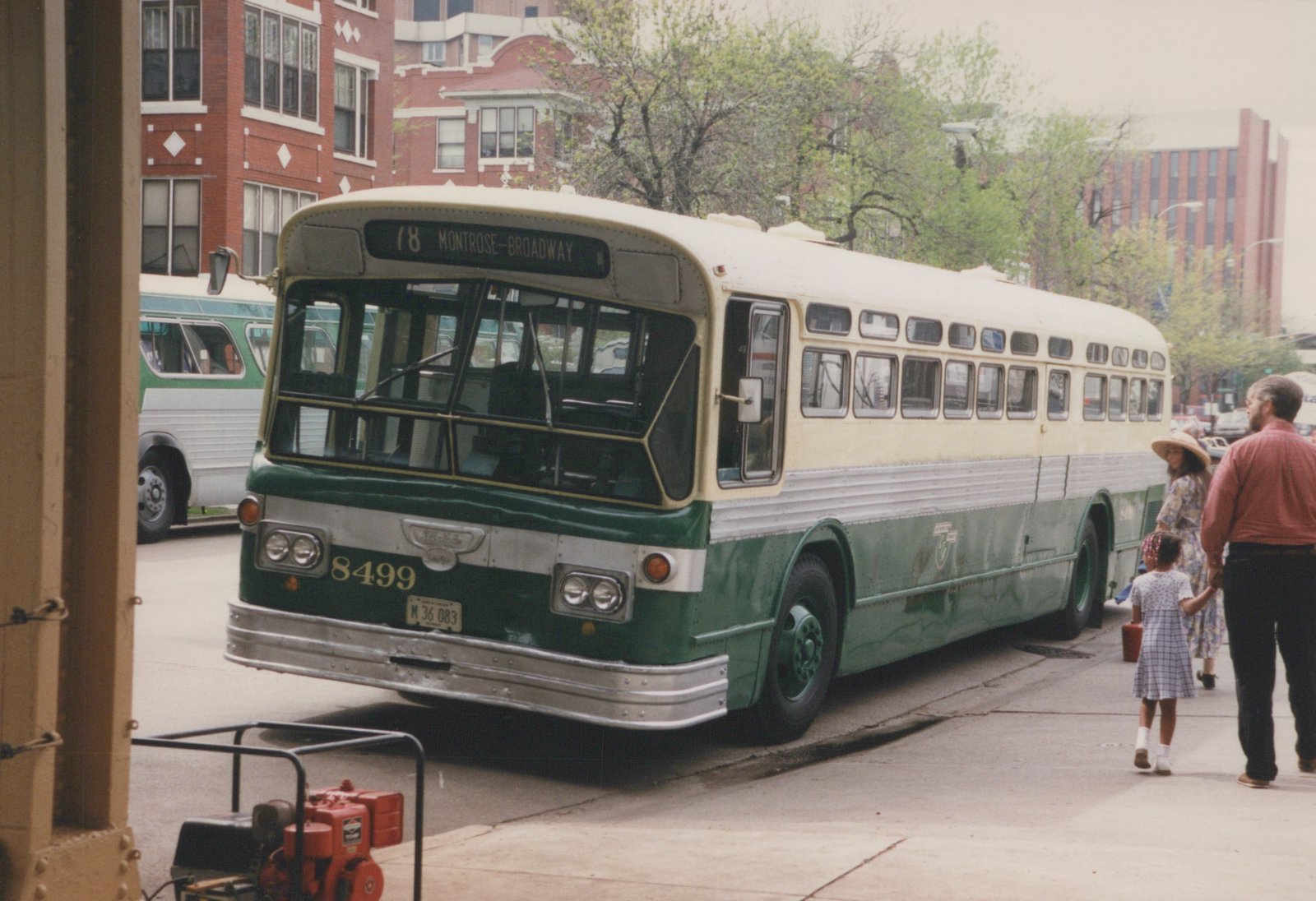 A green and cream CTA bus numbered 8499 is parked on a city street with three people walking nearby; trees with leaves and brick buildings are in the background.