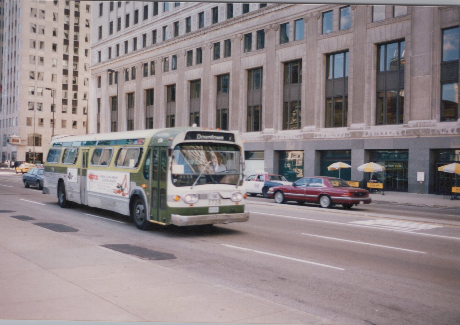 A green and cream Chicago Transit Authority bus, number 7728, drives down a city street in the 1990s, with a 'Downtown' destination sign, surrounded by other cars and tall buildings.