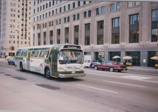 A green and cream Chicago Transit Authority bus, number 7728, drives down a city street in the 1990s, with a 'Downtown' destination sign, surrounded by other cars and tall buildings.