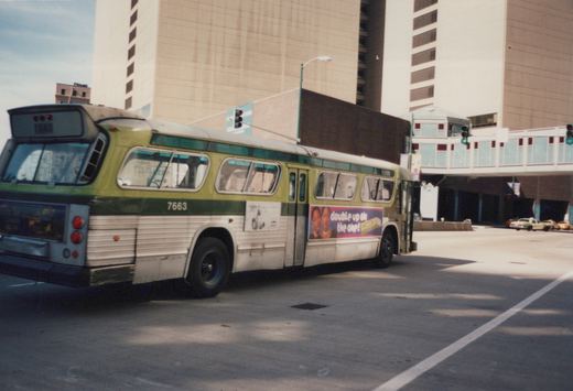 A green and cream CTA bus, number 7663, is parked on a city street with tall buildings and a pedestrian bridge in the background.