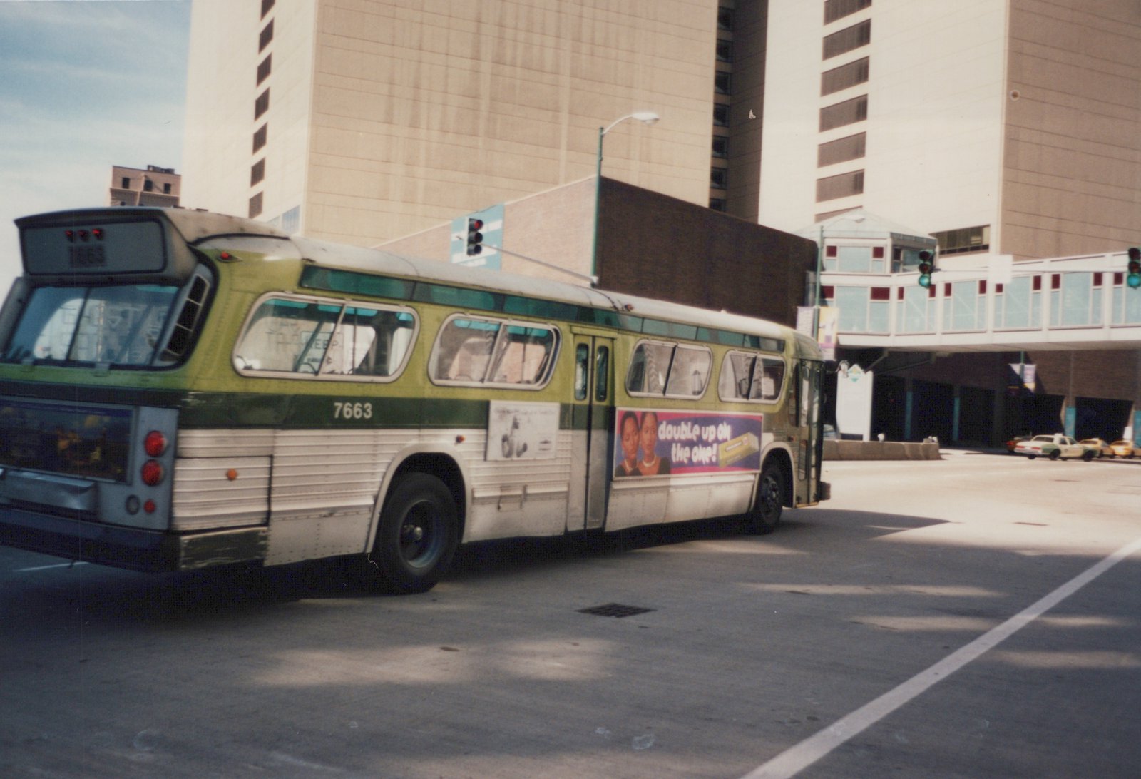 A green and cream CTA bus, number 7663, is parked on a city street with tall buildings and a pedestrian bridge in the background.