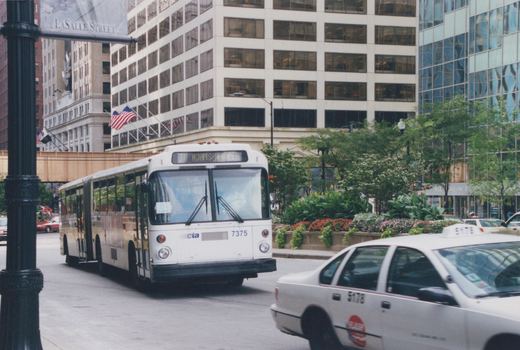 A white Chicago Transit Authority articulated bus numbered 7375 is driving on a city street, with a destination sign reading TO ADAMS/WACKER, and a taxi cab is visible nearby.