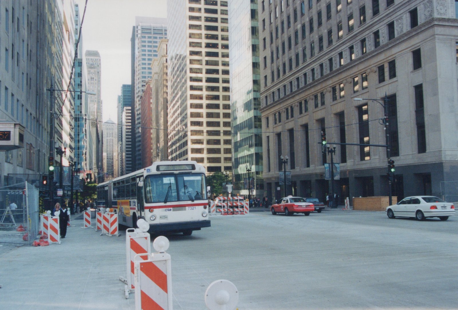 A white and red CTA bus on a city street with tall buildings in the background, construction barriers on the sidewalk, and cars at an intersection.