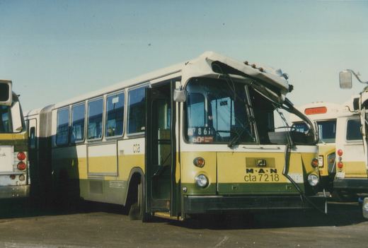A white, yellow and green Chicago Transit Authority bus with the number 7218 on its front, parked outdoors with a damaged roof and windshield frame, and other buses visible nearby.