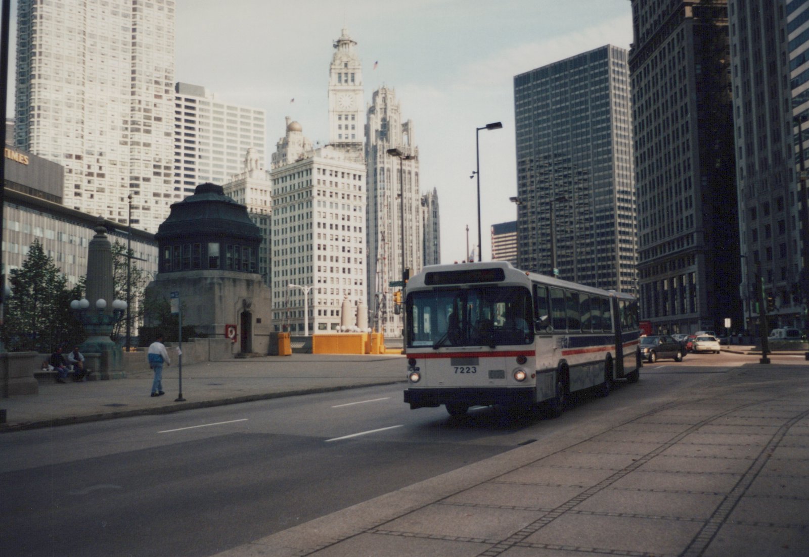 An articulated bus with the number 7223 and CTA lettering drives on a city street, with skyscrapers including the Tribune Tower in the background.