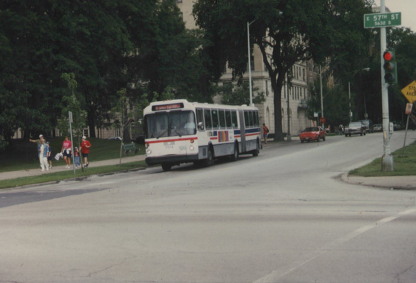 A white CTA articulated bus with the number 7174 is turning a corner on a tree-lined city street, with pedestrians on the sidewalk and cars in the background. A street sign reads 'E 57TH ST'.